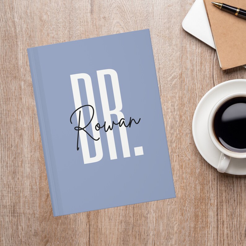 May include: A light blue journal with the name "Rowan" in a handwritten font, and the initials "DR." in a bold, sans-serif font. The journal is on a wooden surface, next to a cup of coffee and a notebook.