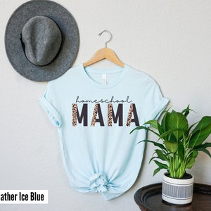 Op de afbeelding: Een lichtblauw T-shirt met de tekst "homeschool MAMA" in zwarte letters met een luipaardprint achter de letters. Het shirt hangt aan een houten hanger.