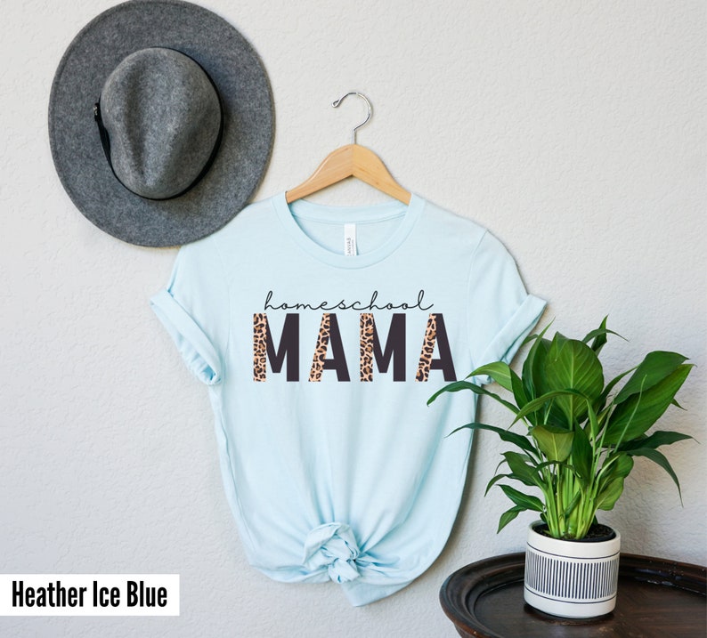 Op de afbeelding: Een lichtblauw T-shirt met de tekst "homeschool MAMA" in zwarte letters met een luipaardprint achter de letters. Het shirt hangt aan een houten hanger.