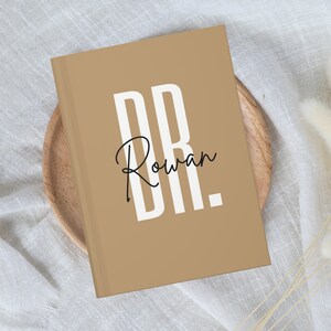 May include: A tan-colored journal with the text "DR. Rowan" in a bold white font. The journal is resting on a wooden plate. The cover has a minimalist design, making it suitable for note-taking or journaling.