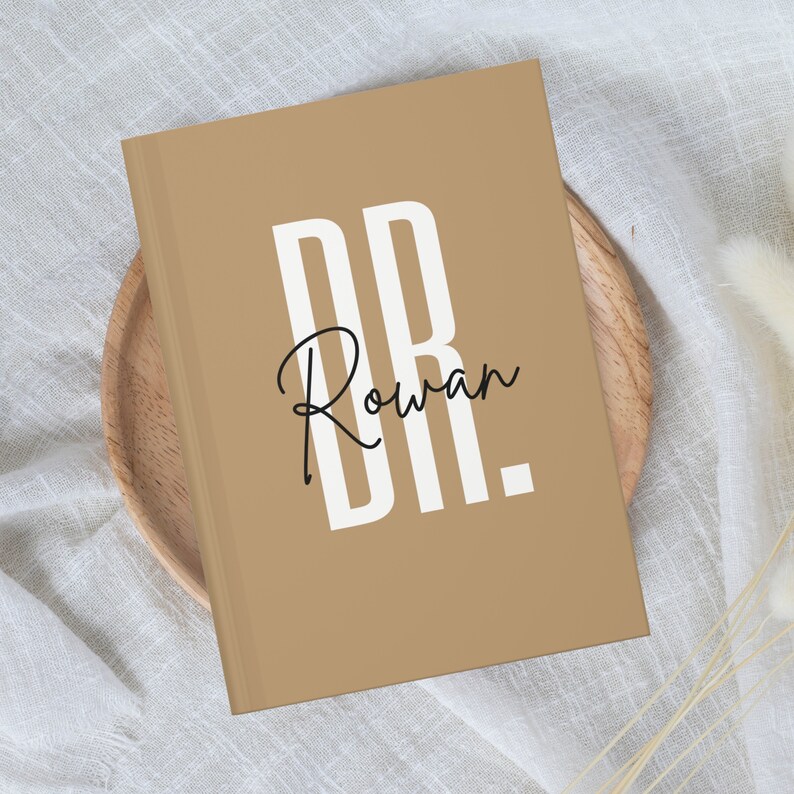 May include: A tan-colored journal with the text "DR. Rowan" in a bold white font. The journal is resting on a wooden plate. The cover has a minimalist design, making it suitable for note-taking or journaling.