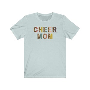 以下が含まれることがあります： 薄い青色のTシャツに、レオパード柄のカラフルな文字で「CHEER MOM」と書かれています。