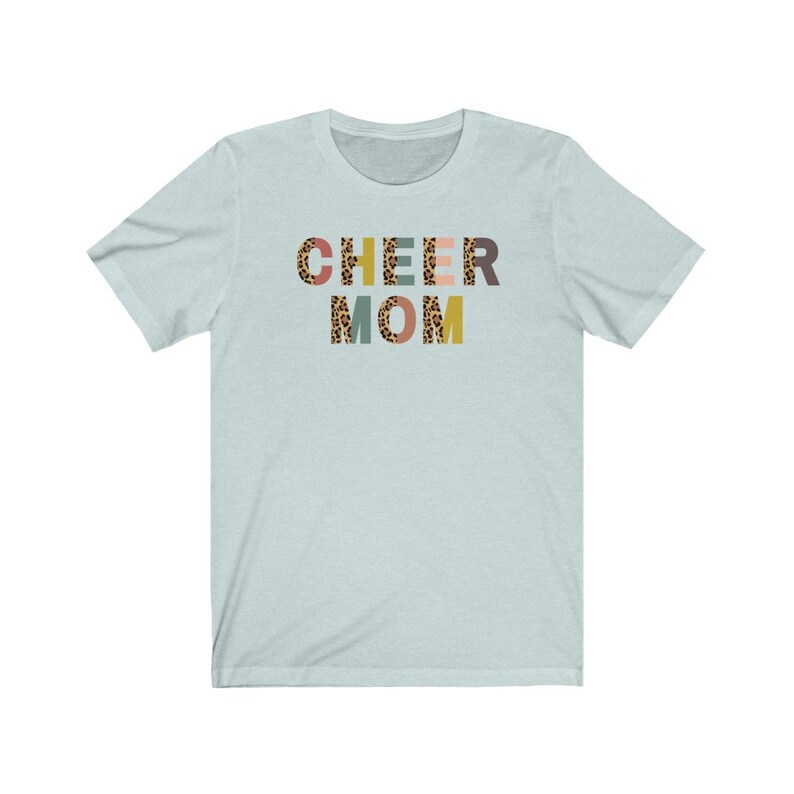 以下が含まれることがあります： 薄い青色のTシャツに、レオパード柄のカラフルな文字で「CHEER MOM」と書かれています。