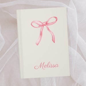 May include: A white journal with a pink watercolor bow and the name "Melissa" written in pink cursive.