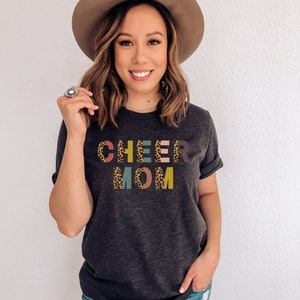 以下が含まれることがあります： ダークヘザーグレーのTシャツに、レオパード柄のカラフルなブロック文字で「CHEER MOM」と書いてあります。