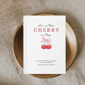 May include: A white book with the text "she is the CHERRY on top" in red and black lettering. The book has a pink ribbon and two red cherries on the front. The text "ADVICE & WISHES FOR THE FUTURE PARENTS HANNAH AND JACKSON" is printed in black at the bottom.