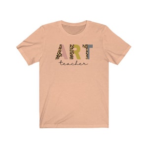 K&ouml;nnte beinhalten: Peachfarbenes T-Shirt mit kurzen &Auml;rmeln mit dem Wort "ART" in bunten Buchstaben mit einem Leopardenmuster. Das Wort "teacher" ist in schwarzer Schrift unter dem Wort "ART" geschrieben.