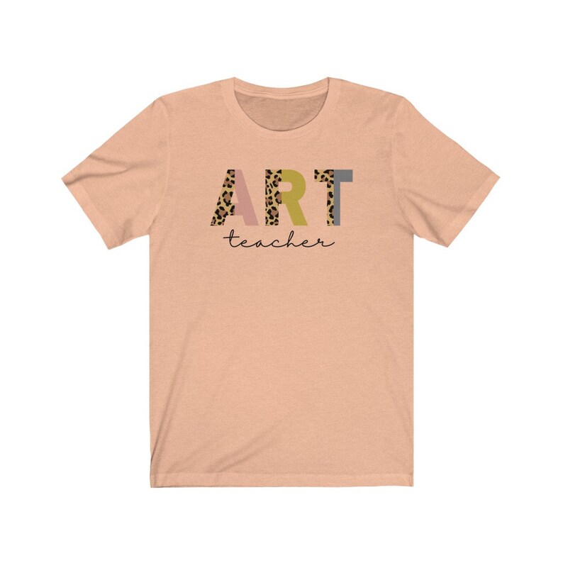 K&ouml;nnte beinhalten: Peachfarbenes T-Shirt mit kurzen &Auml;rmeln mit dem Wort "ART" in bunten Buchstaben mit einem Leopardenmuster. Das Wort "teacher" ist in schwarzer Schrift unter dem Wort "ART" geschrieben.