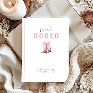 Puede incluir: Un libro blanco con la inscripción "first RODEO" en rosa. Debajo, una ilustración en acuarela de botas de vaquero marrones con un lazo rosa. El libro está personalizado con un nombre y una fecha.