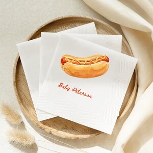 May include: A stack of white paper napkins featuring a watercolor illustration of a hot dog with yellow mustard and the red text Baby Peterson printed below the graphic. The napkins have a textured border and are square in shape.