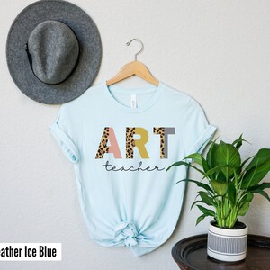 K&ouml;nnte beinhalten: Ein hellblaues T-Shirt mit dem Wort "ART" in bunten Buchstaben mit einem Leopardenmuster. Das Wort "teacher" ist in schwarzer Schrift unter den Buchstaben geschrieben.