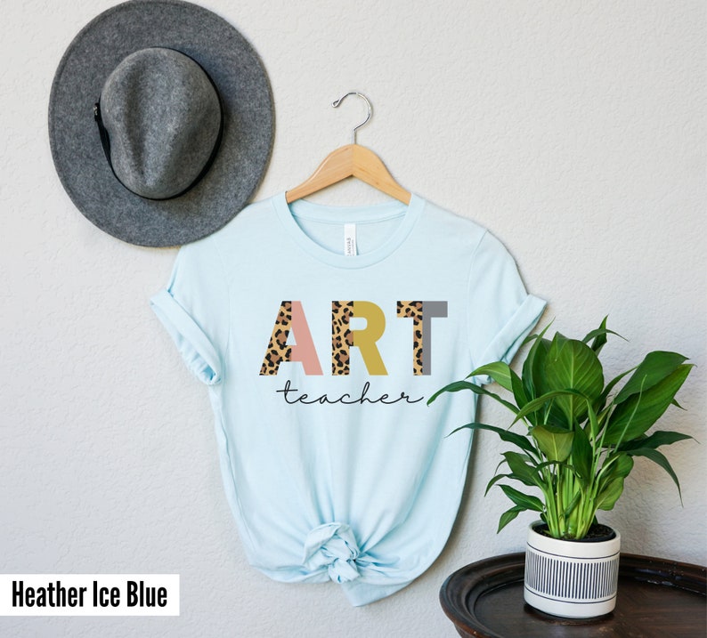 K&ouml;nnte beinhalten: Ein hellblaues T-Shirt mit dem Wort "ART" in bunten Buchstaben mit einem Leopardenmuster. Das Wort "teacher" ist in schwarzer Schrift unter den Buchstaben geschrieben.