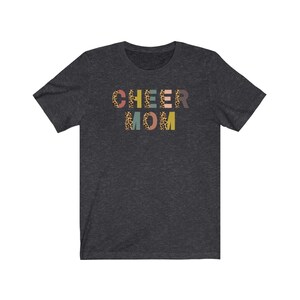 以下が含まれることがあります： レオパード柄のカラフルな文字で「CHEER MOM」と書かれたダークヘザーグレーのTシャツです。