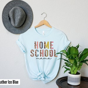 May include: A light blue t-shirt with the text "HOME SCHOOL mama" in colorful letters with a leopard print pattern. The shirt is hanging on a wooden hanger.