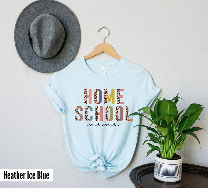 May include: A light blue t-shirt with the text "HOME SCHOOL mama" in colorful letters with a leopard print pattern. The shirt is hanging on a wooden hanger.