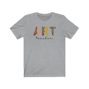 K&ouml;nnte beinhalten: Ein heidefarbenes T-Shirt mit dem Wort "ART" in bunten Buchstaben mit Leopardenmuster. Das Wort "teacher" ist in schwarzer Schrift unter dem Wort "ART" geschrieben.
