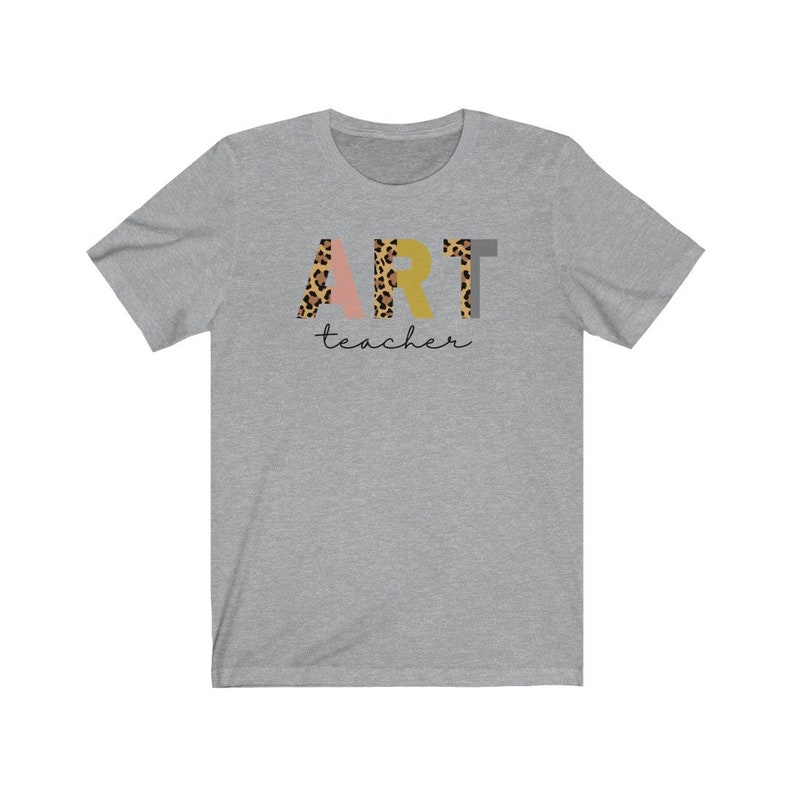 K&ouml;nnte beinhalten: Ein heidefarbenes T-Shirt mit dem Wort "ART" in bunten Buchstaben mit Leopardenmuster. Das Wort "teacher" ist in schwarzer Schrift unter dem Wort "ART" geschrieben.