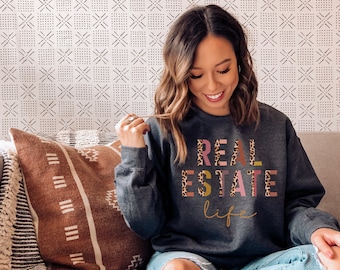 May include: A gray sweatshirt with the text "REAL ESTATE life" in colorful letters with a leopard print pattern. The letters are in different colors, including pink, yellow, and green.
