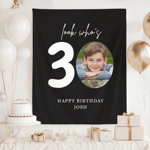 May include: Black birthday banner with the text "look who's 30" and "Happy Birthday Josh". The number 30 is in large white font. An oval photo of a young person is in the center. White and gold balloons are in the background.