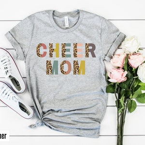 以下が含まれることがあります： レオパード柄のカラフルな文字で「CHEER MOM」と書かれたグレーのTシャツ。文字はブロック体です。