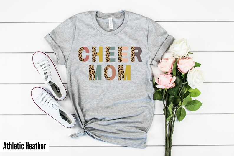 以下が含まれることがあります： レオパード柄のカラフルな文字で「CHEER MOM」と書かれたグレーのTシャツ。文字はブロック体です。