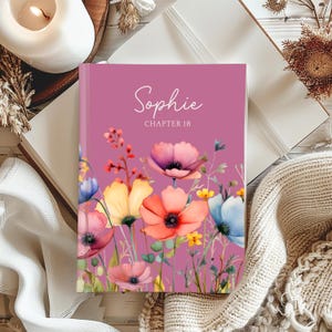 May include: A hardcover journal with a floral design. The cover is a shade of purple with the name "Sophie" and "Chapter 18" in white script. The floral design features watercolor-style flowers in shades of pink, orange, yellow, and blue.