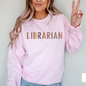 Peut inclure: Un sweat-shirt rose p&acirc;le avec le mot "LIBRARIAN" imprim&eacute; sur le devant en lettres color&eacute;es avec un motif l&eacute;opard.