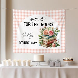 May include: A pink and white gingham backdrop with the text "one FOR THE BOOKS" and "Sally's 1ST BIRTHDAY". The backdrop features a floral illustration of books and roses. A table is set with a cake, cupcakes, and gifts.