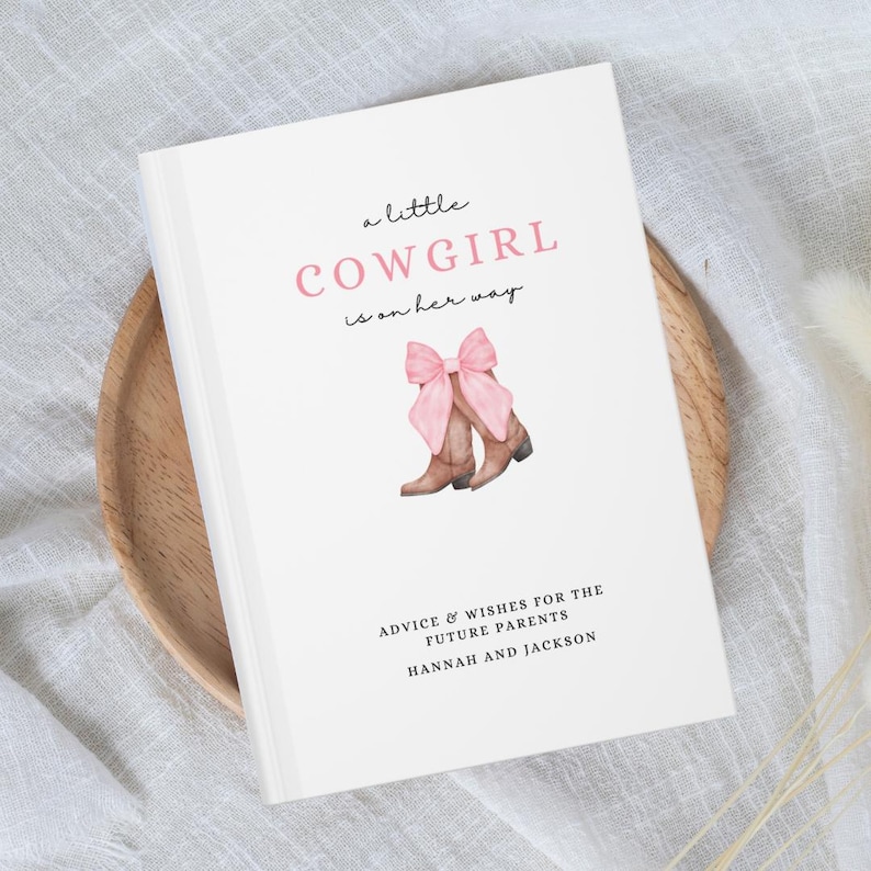 Puede incluir: Un libro blanco con el texto "a little COWGIRL is on her way" en letras rosas y negras. El libro tiene una ilustraci&oacute;n de acuarela de botas de vaquero marrones con lazos rosas en la parte superior. El texto "ADVICE & WISHES FOR THE FUTURE PARENTS HANNAH AND JACKSON" est&aacute; impreso en negro en la parte inferior del libro.