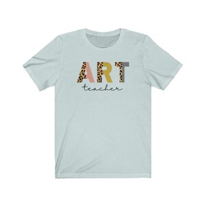 K&ouml;nnte beinhalten: Hellblaues T-Shirt mit dem Wort "ART" in bunten Buchstaben mit Leopardenmuster. Das Wort "teacher" ist in schwarzer Schrift unter den Buchstaben geschrieben.
