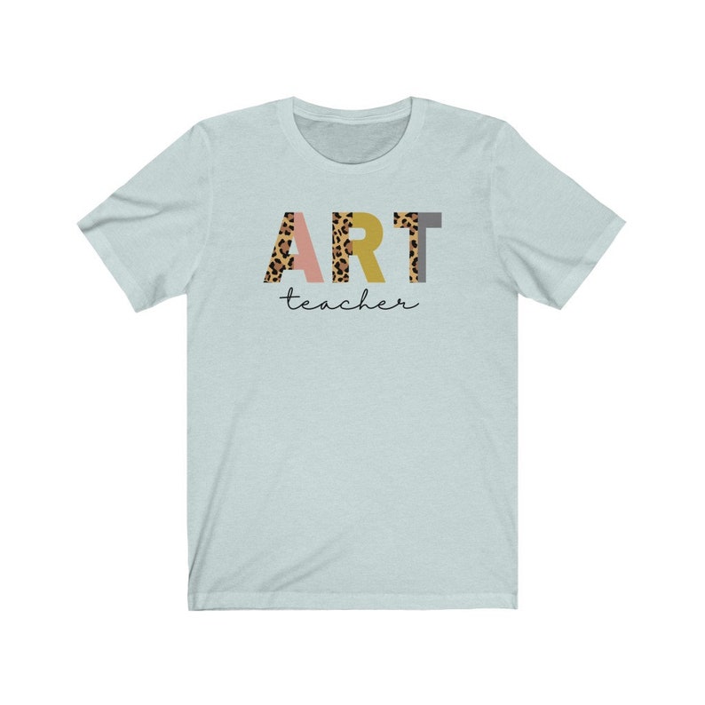K&ouml;nnte beinhalten: Hellblaues T-Shirt mit dem Wort "ART" in bunten Buchstaben mit Leopardenmuster. Das Wort "teacher" ist in schwarzer Schrift unter den Buchstaben geschrieben.
