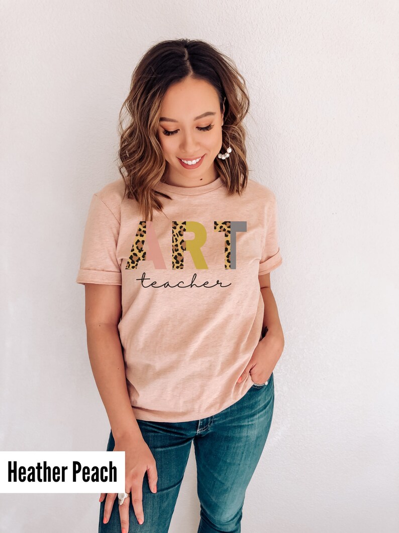 K&ouml;nnte beinhalten: Ein T-Shirt in der Farbe Heidepfirsich mit dem Wort "ART" in Rosa, Gelb und Grau mit einem Leopardenmuster. Das Wort "teacher" ist darunter in Kursivschrift geschrieben.