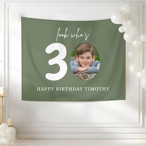 May include: Olive green birthday banner with the text "look who's 3" and "HAPPY BIRTHDAY TIMOTHY" in white. A photo of a young boy is in the center. White balloons are in the background.