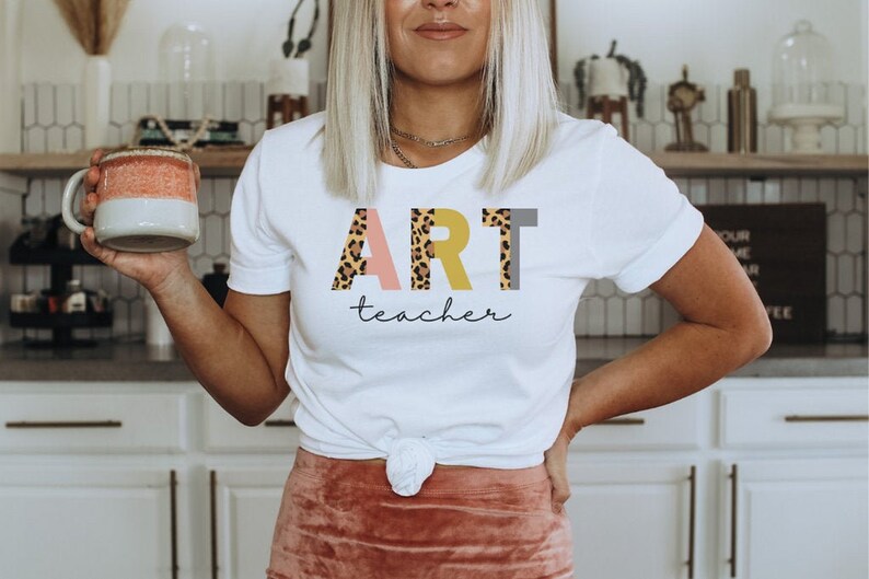 K&ouml;nnte beinhalten: Wei&szlig;es T-Shirt mit dem Wort "ART" in Leopard-Print, rosa, gelben und grauen Buchstaben. Der Text "teacher" ist darunter in Kursivschrift geschrieben.