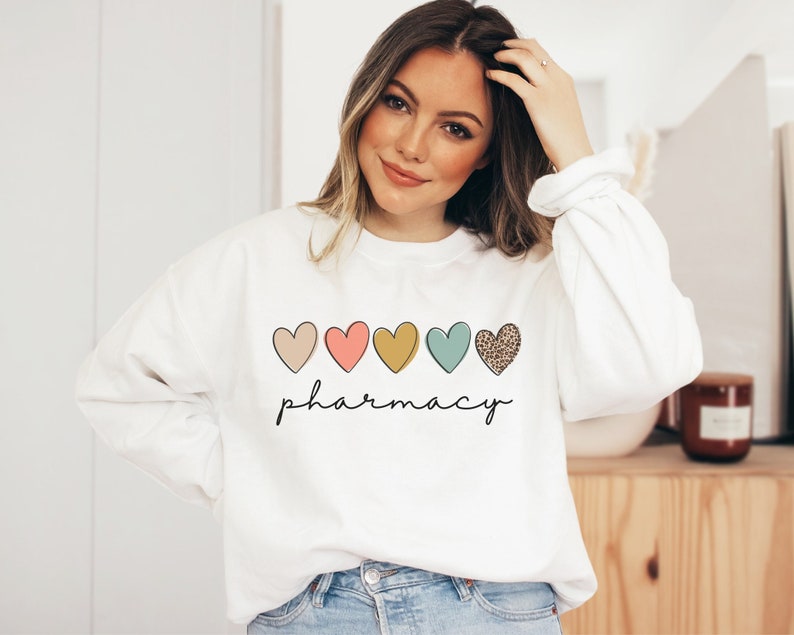 May include: White sweatshirt with a row of five colorful hearts and the word "pharmacy" written below. The hearts are pink, peach, yellow, teal, and brown with a leopard print.