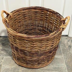 May include: A large, round, open-top basket woven from natural brown willow branches. The basket features a sturdy, thick rim and two integrated, braided side handles. The weave pattern is consistent throughout, displaying varied shades of brown.