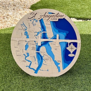 May include: A round wooden map of St. Augustine, Florida, with a layered design showing the city's layout and waterways. The map features blue water details, a bridge, and the city's name in script. A compass rose is visible.