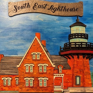 Puede incluir: Un letrero de madera que representa el faro de South East. El faro es de ladrillo rojo con una torre de luz negra y verde. El letrero de arriba dice "South East Lighthouse". El fondo está pintado de azul. Es un objeto decorativo.