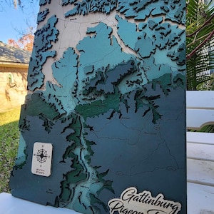 May include: A wooden map of Gatlinburg and Pigeon Forge, Tennessee. The map is layered and has a blue and green color scheme. The map is mounted on a wooden frame.
