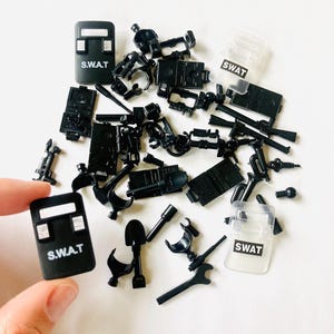 May include: A collection of black toy SWAT team accessories, including shields with "S.W.A.T" text, weapons, and other tactical gear. The items are scattered on a white surface.