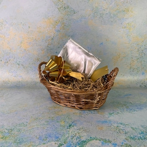 May include: An oval-shaped wicker basket with a gold bow, shredded paper, and a clear cellophane bag. A gold gift tag is also inside the basket. The basket has handles and is set against a textured background.