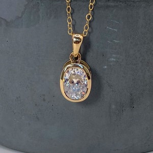 May include: A gold-colored necklace with an oval-shaped, clear gemstone pendant. The pendant is set in a gold bezel and hangs from a delicate gold chain. The necklace is displayed on a gray, cylindrical surface.