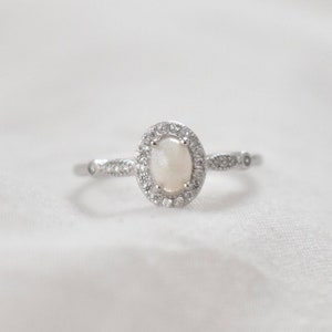 May include: A silver ring with a white oval gemstone surrounded by smaller clear gemstones.