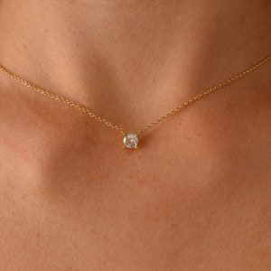May include: A delicate gold necklace featuring a small, round, clear gemstone pendant. The pendant is centered on a fine gold chain, creating a minimalist and elegant design. The necklace is displayed on a person's chest.