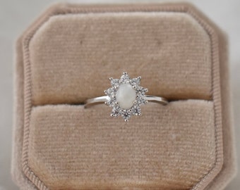 May include: A silver ring with a white oval stone surrounded by smaller white stones in a starburst design. The ring is displayed in a beige velvet ring box.