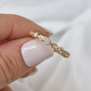 Può includere: Un anello dorato con pietre chiare e a forma di pera di colore chiaro. L'anello è tenuto su uno sfondo bianco, mostrando il suo design delicato e la sua lucentezza. La fascia dell'anello è sottile e le pietre sono molto vicine.