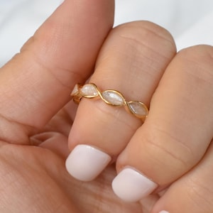 Pode incluir: Um anel dourado com um padrão repetitivo de pedras ovais esbranquiçadas. O anel é usado em um dedo, com a mão e as unhas visíveis. O design do anel é um laço contínuo de ouro e pedras entrelaçadas.