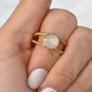 May include: A gold ring featuring a circular, pale-toned stone. The band has a twisted rope pattern, and the stone is secured by a slender gold rim. The ring is displayed on a hand with light-coloured nail polish.