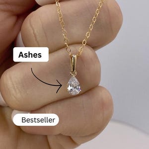 Peut inclure: Un collier doré avec un pendentif en forme de poire en pierre précieuse transparente. Le collier est tenu par une personne. Le mot "Ashes" est écrit au-dessus du pendentif et le mot "Bestseller" est écrit en dessous.