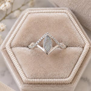 May include: A silver chevron-shaped ring featuring a central marquise-cut grey stone and a band accented with small clear stones. The ring is displayed in a beige velvet hexagonal box, showcasing a pointed design that frames the center stone.
